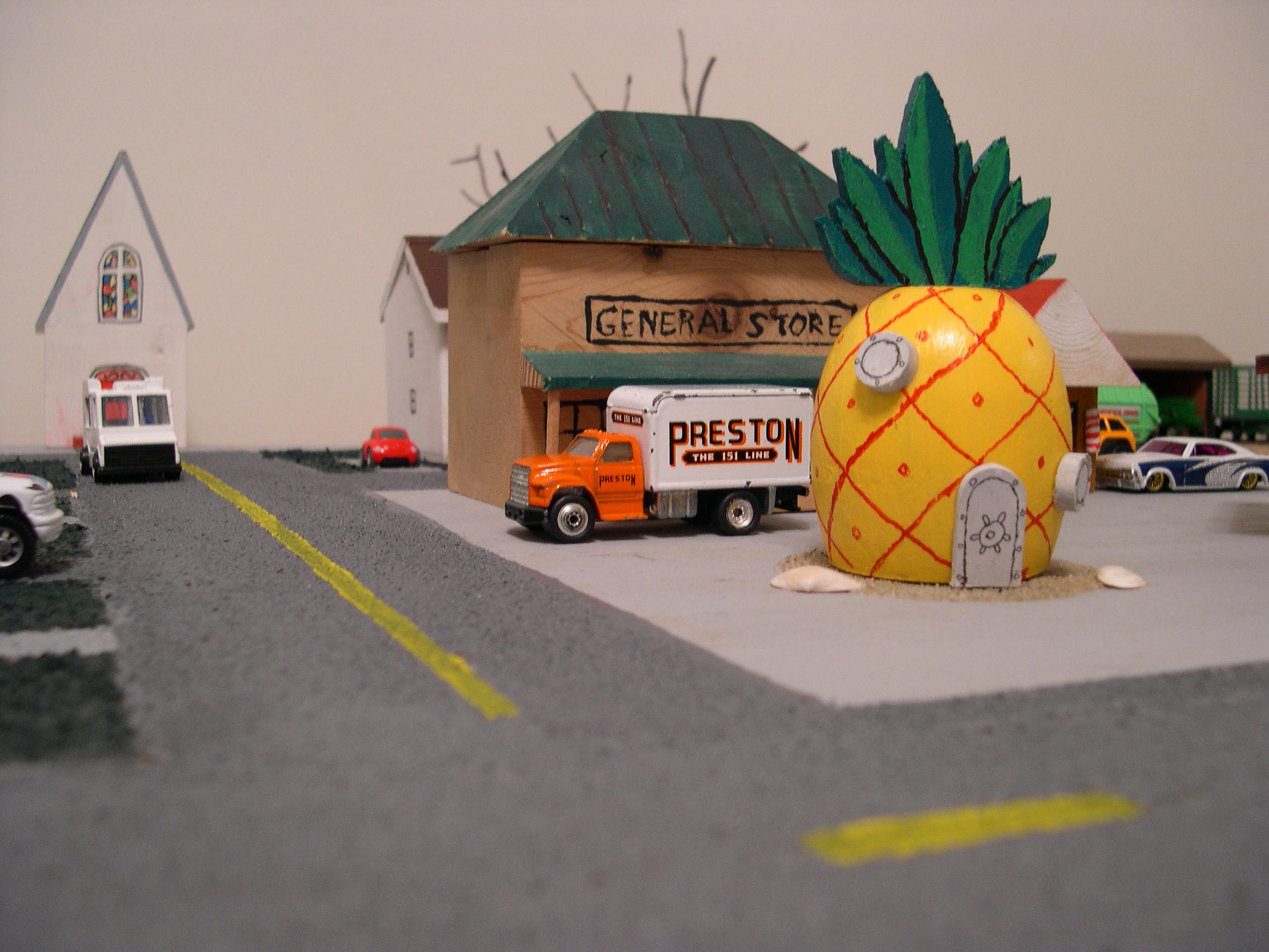 Wide neighborhood view showing SpongeBob's pineapple house next to a vintage general store with toy delivery truck and white church building on a miniature street scene